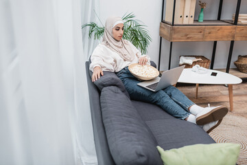 Fototapeta na wymiar full length view of arabian woman in hijab sitting on sofa with popcorn and watching film on computer.