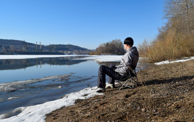 A man is sitting on a folding chair on the bank of an icy river on a spring day