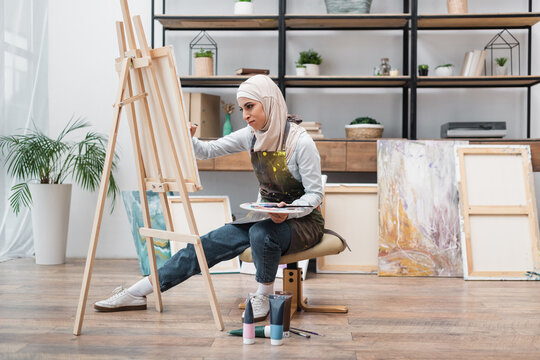 full length view of muslim woman drawing on easel near paint tubes on floor.