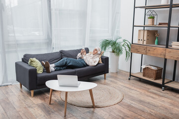happy muslim woman listening music in headphones while lying on couch in living room.