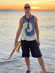 Young male saxophonist stands with his feet in sea water, holds saxophone in his hands. Beautiful sunset on sea, sky. Musician, playing saxophone, dancing, Silhouette. Wearing dark black glasses
