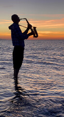 Young male saxophonist stands with his feet in sea water, holds saxophone in his hands, looks at...