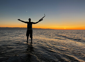 Fototapeta na wymiar Young male saxophonist stands with his feet in sea water, holds saxophone in his hands, looks at sunset. Beautiful sunset on sea, sky. Musician, playing saxophone, dancing, having fun, Silhouette