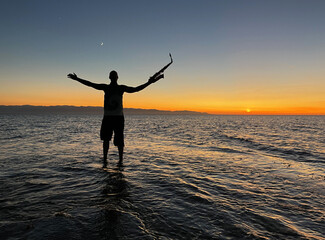 Young male saxophonist stands with his feet in sea water, holds saxophone in his hands, looks at sunset. Beautiful sunset on sea, sky. Musician, playing saxophone, dancing, having fun, Silhouette