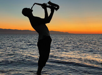 Young male saxophonist stands with his feet in sea water, holds saxophone in his hands, looks at sunset. Beautiful sunset on sea, sky. Musician, playing saxophone, dancing, having fun, Silhouette