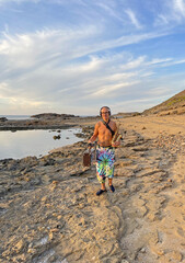 A musician saxophonist with a naked torso in bright multi-colored wide pants and headphones walks along the seashore with a saxophone suitcase