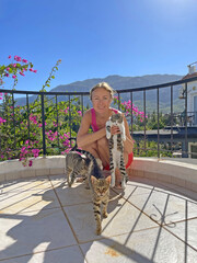 Young beautiful girl smiles, holds Street cats in her arms, in summer in nature in Cyprus, balcony against background of blue sky, mountains. Love of animals, care, tenderness, friendship, affection