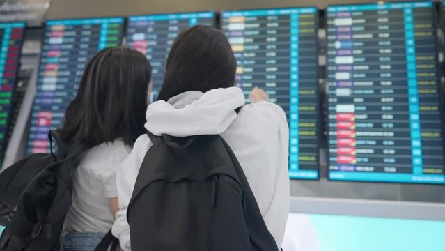 Rear view of Two asian females passengers standing and looking at airport time table board, check in for flight, travel during epidemic adapt to new normal lifestyle, safe travel insurance