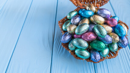Multicolored chocolate eggs in a wicker basket on a blue wooden table on easter day with empty space for text on the right