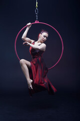 Circus performer woman in red dress doing tricks on red Lyra isolated on black background.
