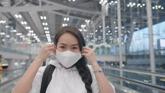 Happy attractive asian tan skin female traveler wearing medical face mask on moving walkalator inside modern glass airport terminal, self respiratory system protection, passenger fly through, 