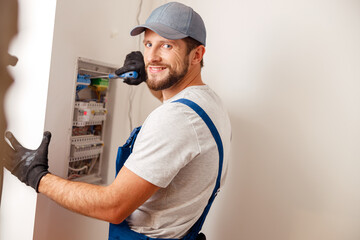 Electrical technician in uniform smiling at camera, using screwdriver while checking fuses in a...