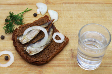 Sprats on a slice of black bread sprinkled with onion rings and herbs, with a glass of vodka on a serving board