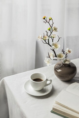 The atmosphere of a romantic spring morning. Blooming branches in a vase, a book and a cup of coffee on a white table in a Provence style garden. Home interior. Life style