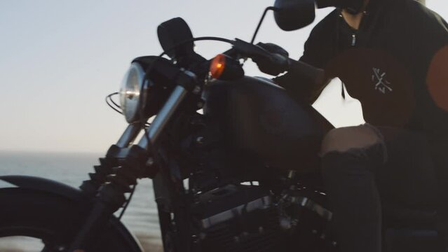 Man, wearing helmet and tracksuit, ride black bike on a coastal highway at sunset. Close-up shot of a motorcyclist on summer journey within beautiful landscape and sea behind. High quality 4k footage