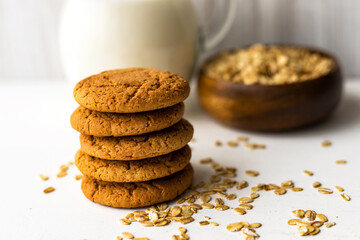 Heap oatmeal cookies and oat cereal flakes bowl on white table on milk glass jug background