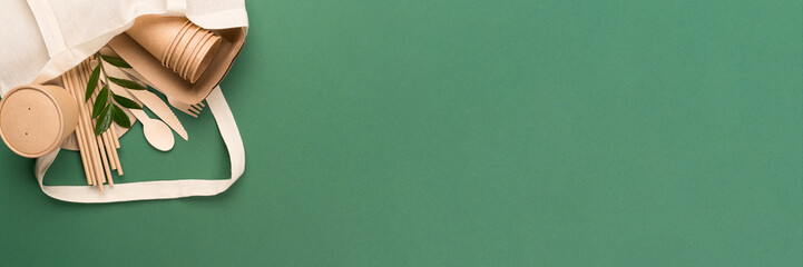 Banner with eco-friendly paper tableware - kraft paper food cups and containers in cotton bag on green background with copy space. Street food take away paper utensils - cups, plates, drinking straws
