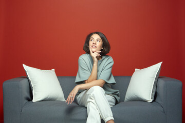Thoughtful young beautiful girl dreaming sitting on sofa. Pensive woman touching chin looking up,...