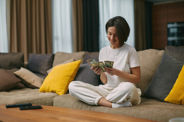 Happy young woman counting cash, enjoying money win, big profit, salary in payday sitting on sofa...