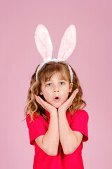 Obraz na płótnie Canvas Adorable surprised little girl with long curly hair in Easter bunny ears touching cheeks and looking at camera with open-eyes, against pink background in studio, vertical orientation