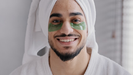 Funny portrait male face smiling indian arabian man wearing bath towel on head glue sticky hydrogel eye-patches cosmetic procedures skin care guy looking at camera with collagen patches under eyes