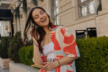Image of young beautiful brownhaired posing relaxed against backdrop on street. Caucasian woman with closed eyes listens to music with headphones using mobile phone. Emotion concept, facial expression