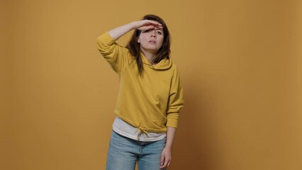 Curious woman looking into the distance far away searching for something with palm over forhead in studio. Young person scouting the horizon seeking for someone over yellow background.