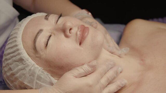 Young hypnotic face woman receiving anti-ageing facial massage in spa salon relax. Wellness body skin care face beauty treatment, rejuvenation procedure getting head face massage. Black background