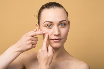 Pretty caucasian girl squeezing a pimple on her cheek isolated on a beige studio background. Skin...