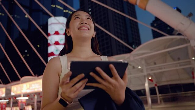 at night,young Asian woman happily plays with the tablet .She looks forward with hopes and dreams, a technology device of today in the middle of an urban society with buildings a in the backdrop.
