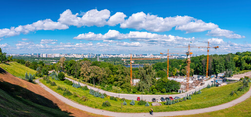 Scenic view of construction cranes in park with cityscape on sunny day against cloudy sky, Industrial cranes at construction site in park