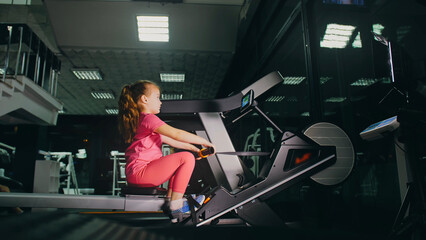 Fototapeta na wymiar Fit Athletic Baby Girl Work out on Rowing Machine, Doing Her Fitness Exercise. Children Activity Training in Modern Gym. Sports Child Workout in Exercising on Row Machine Fitness Center.