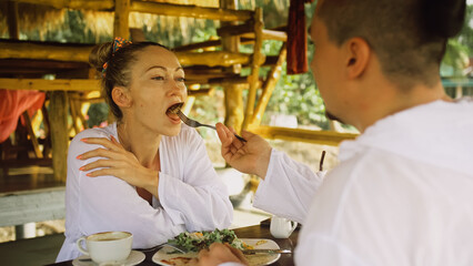Attractive young man and woman eat and drink tasty beverages spending time in traditional local floating cafe on water. Loving happy couple having breakfast outdoors, feed each other. Tropical view