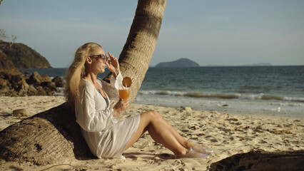 Fototapeta na wymiar Seductive woman sitting in white dress sunglasses, drinking pineapple cocktail Pina Colada. Palm tree, beach, sea waves at sunset. Concept rest tropical resort traveling tourism happy summer holidays