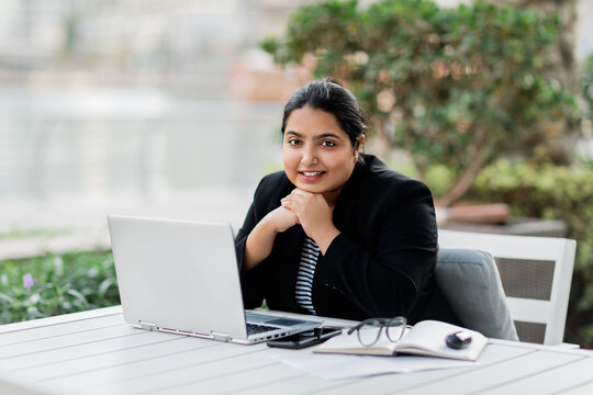 A young and confident Indian Asian woman smiles while sitting and working on her laptop in a stylish cafe on the street on a summer day.