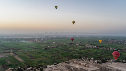 Balloons fly over the Nile Valley at dawn. The sky is bluish-pink. Below are green cultivated fields and an archaeological excavation area. Luxor. Egypt