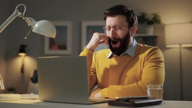 Man yawns, boredom emotion. Tired sleepy bored man with glasses in home office looking at computer screen and yawns for long time