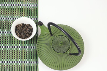 Green iron tea pot with loose tea leaves and bamboo mat on white background.