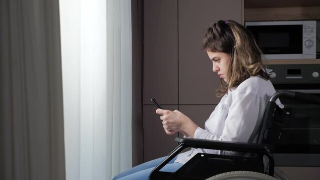 Disabled woman dressed in white shirt tries to type message in smartphone sitting in wheelchair near window behind curtain at home side view.