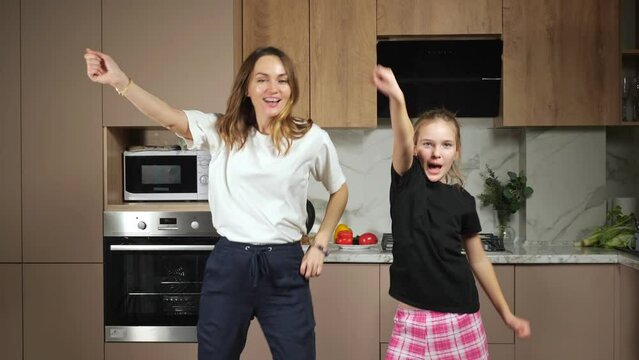 Cheerful long haired mother and teen daughter in black and white t-shirts have fun dancing in kitchen against marble countertop at home.