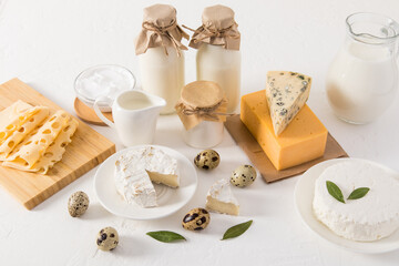 a set of fresh natural products - milk, cottage cheese, cheese, sour cream and eggs in various dishes on a white table against a white brick wall.
