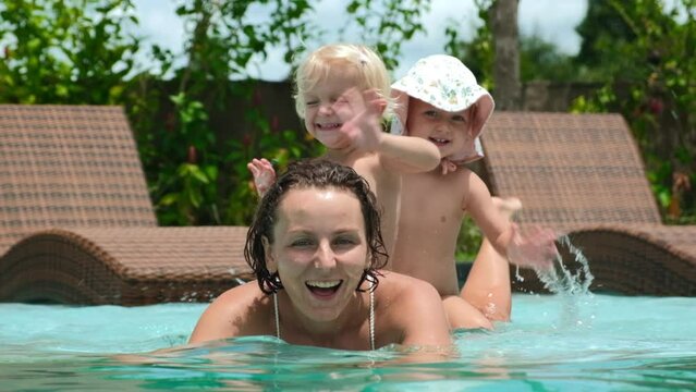 Cheerful family of young mother and toddler twin sisters swimming in pool, waving hello to the camera