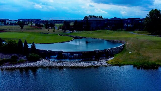 aerial fly into manmade golf course pond waterfalls tier reflecting the cloudy skyline of the fairway luxury homes surrounded by fences manicured gardens pebbles stones in hometown Jagare Summerside