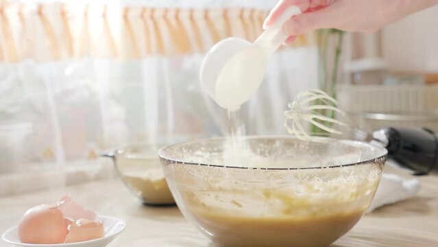 Close-up of a woman's hand pouring flour with a measuring spoon into a glass bowl with dough. Cooking homemade food concept.