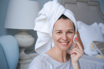 Close-up head shot smiling beautiful woman wrapped in towel applying toner with a cotton swab on her face skin after a bath. Happy young lady doing morning skin care after shower.