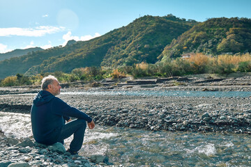 Fototapeta na wymiar Side view of pensive bearded mature man sitting on riverside of mountain river, contemplating nature. Concept of leisure activities,tourism, lifestyle e nature.