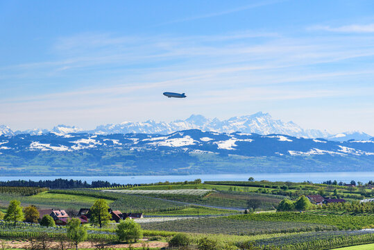 Luftschiff über der Obstbau-Landschaft am Bodensee mit Blick auf den See und die verschneite Alpenkette.
