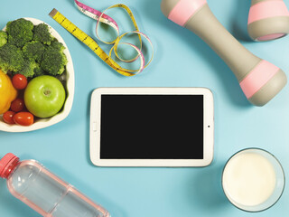black blank screen computer tablet , vegetables and fruit in heart shape plate, bottle of water, measure tape,  pink dumbbells and milk on blue background.