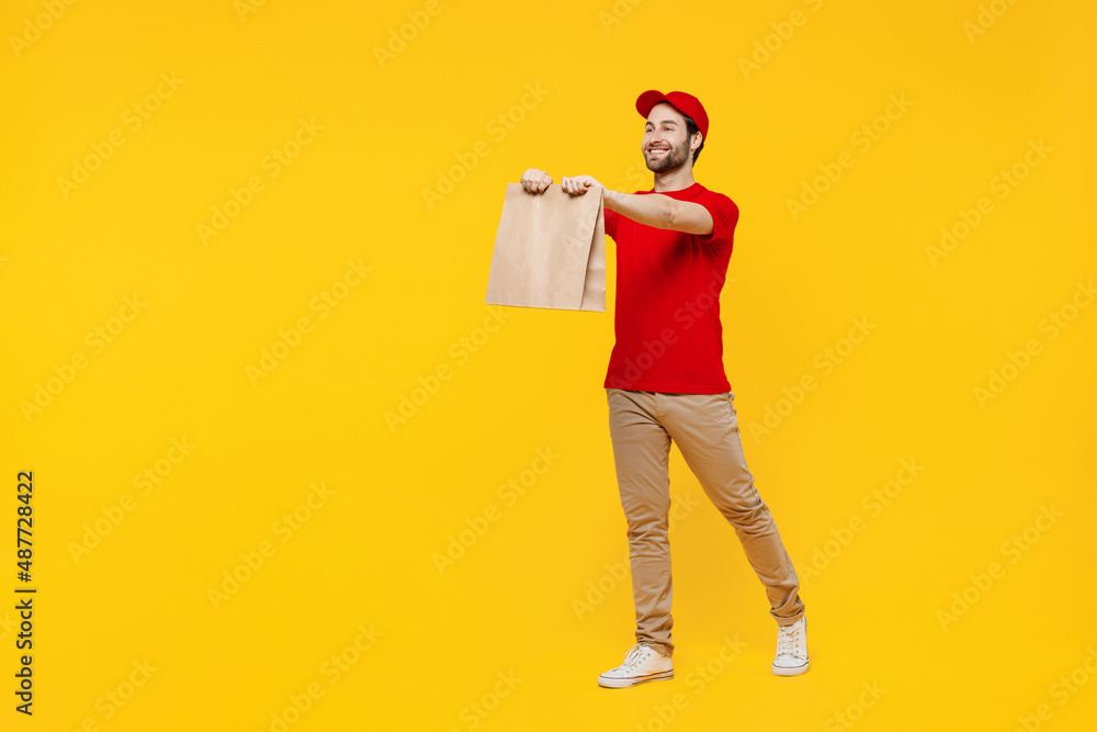 Wall mural Full body fun delivery guy employee man in red cap T-shirt uniform workwear work as dealer courier give brown clear blank craft paper takeaway bag mock up isolated on plain yellow background studio