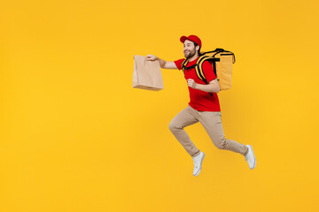 Full body delivery employee man in red cap T-shirt uniform work as dealer courier jump high hold cardboard craft paper takeaway bag run thermal food bag backpack isolated on plain yellow background